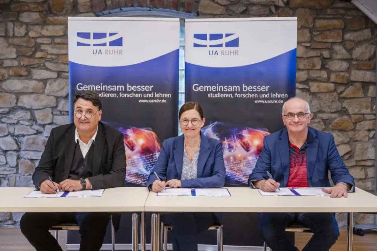 Rektor Paul, Rektorin Albert und Rektor Bayer unterschreiben ein Schriftstück, im Hintergrund ein Banner der UA Ruhr