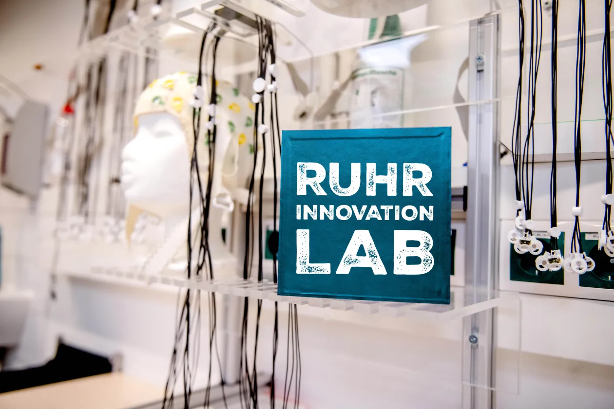 Ein Labor mit EEG-Kappe und Elektroden, darin ein türkisfarbener Würfel mit dem Schriftzug "Ruhr Innovation Lab"