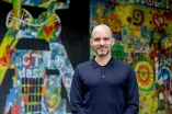 Mann im blauen Pullover vor einer bunt bemalten Wand